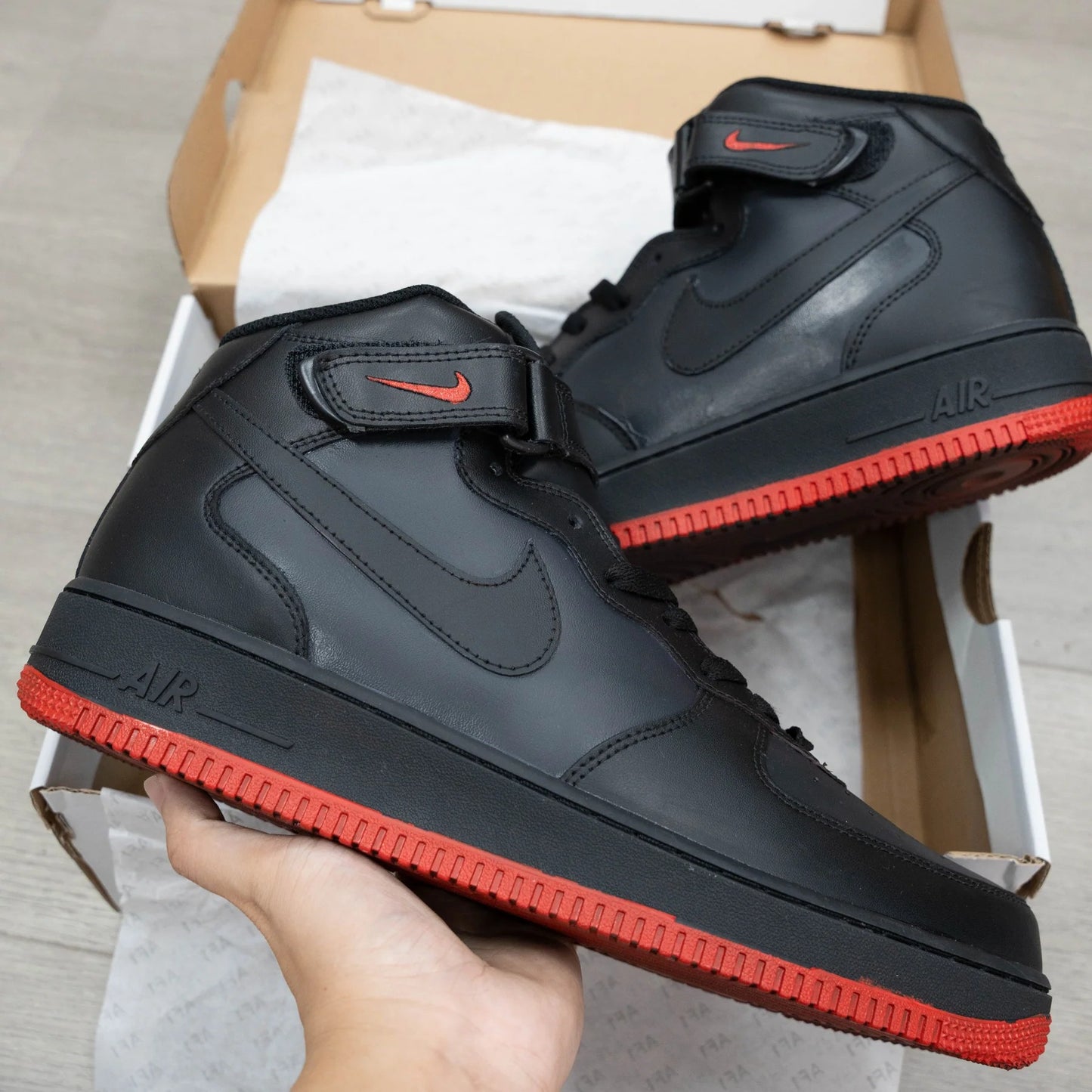 Black high-top sneakers with red soles held in front of a cardboard box.